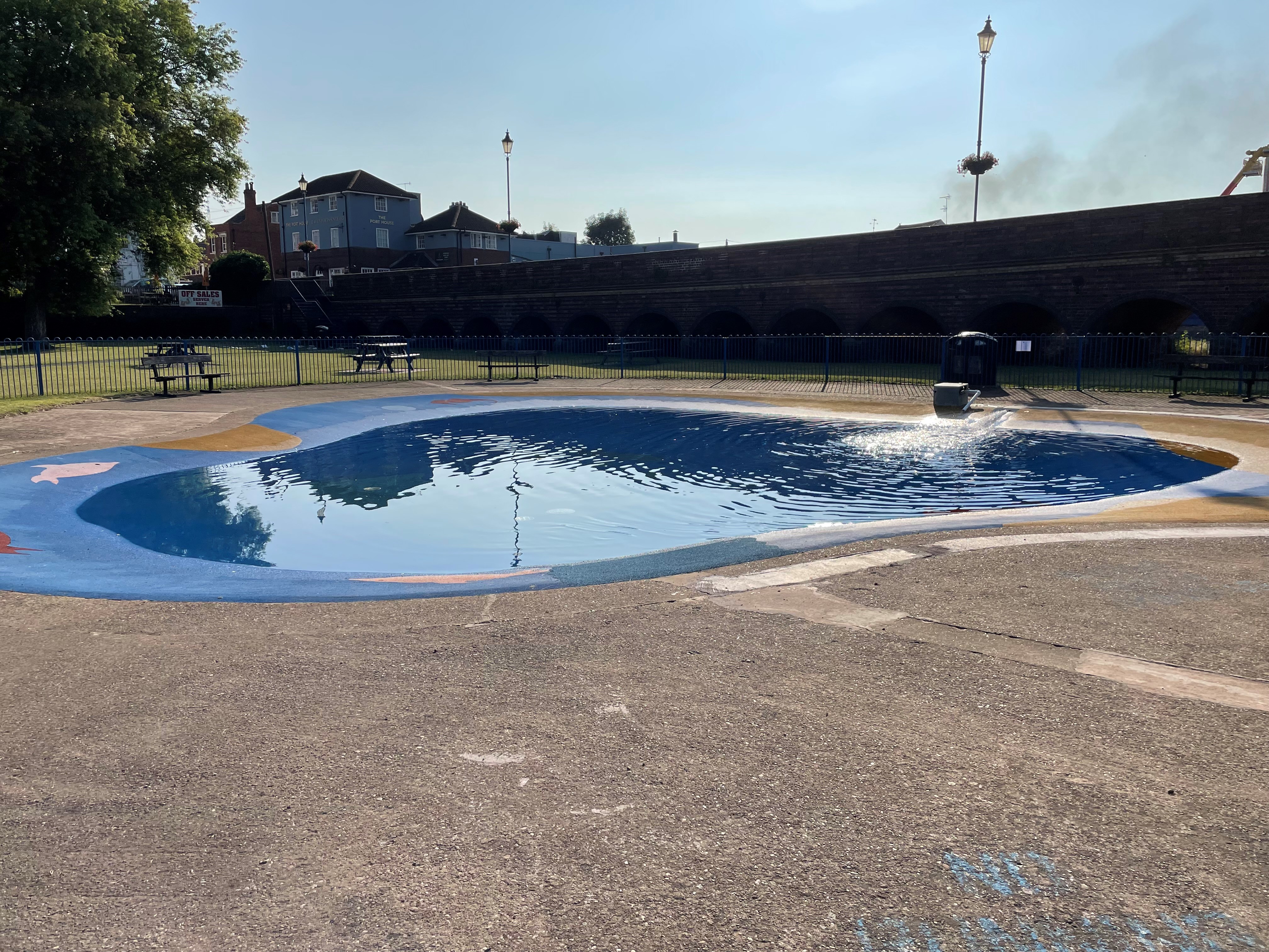 large concrete paddling pool in park with raised road and buildings in distance