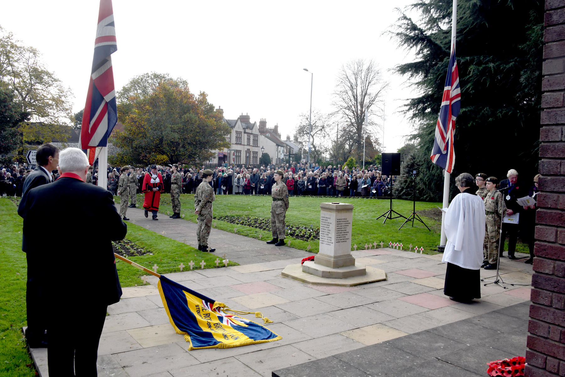 flag bearers with lowered flags stand either side of vicar in front of war memorial reading to gathered crowd