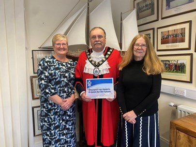3 people smiling at camera, central man in mayoral robes holding printed document