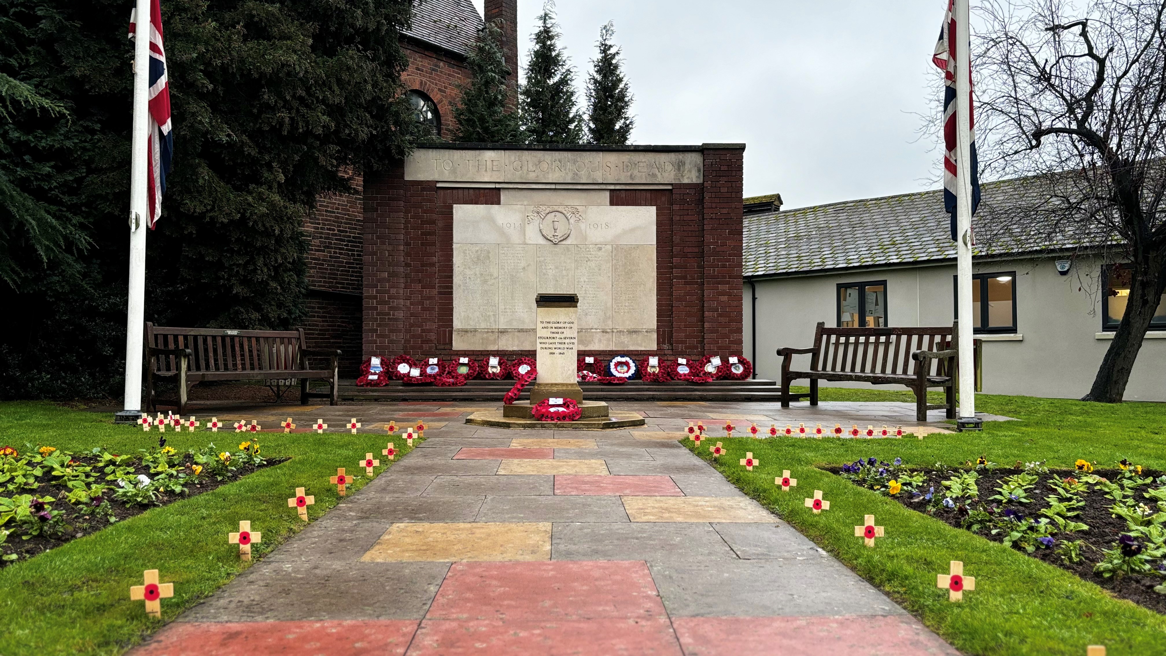 a large brick built war memorial with a smaller pillar in front. poppies on crosses line the path to the memorials, poppy wreaths sit along the bottom of the memorial